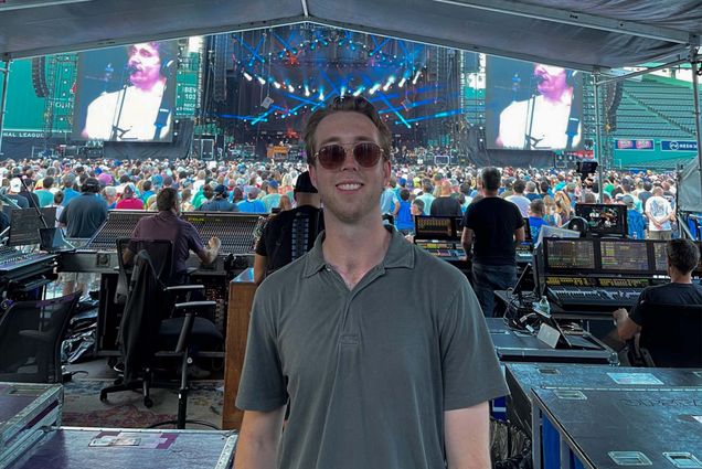 Photo: A college student with short hair wearing sunglasses and a polo shirt stands in front of an audio booth at the back of a crowded concert.
