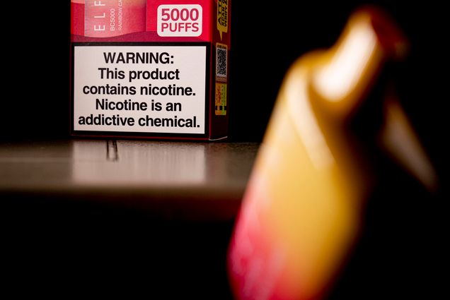 Photo: An Elf Bar disposable vaping pod device is displayed in the foreground, blurred. Behind the vape stands the packaging with a big warning label at the bottom.