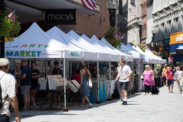 Photo: A bright sunny day with people walking about on a street lined with white tents for a market.