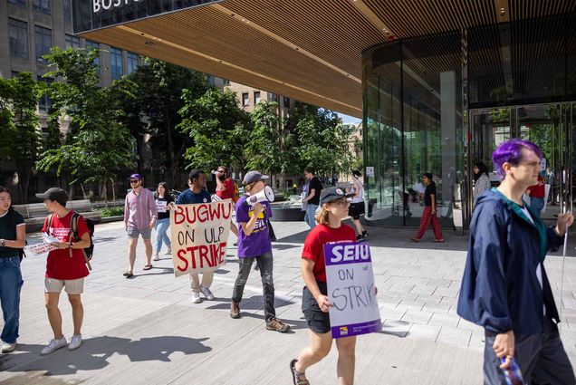 Photo: A group of people participating in a strike or protest outside a building with a modern glass facade. The protestors are holding signs, including one that reads "BUGWU ON ULP STRIKE" and another that says "SEIU ON STRIKE." Many of the participants are wearing shirts with logos, and some are carrying megaphones. The setting appears to be in an urban environment, with trees and buildings visible in the background. The scene depicts a peaceful demonstration with people walking in a line.