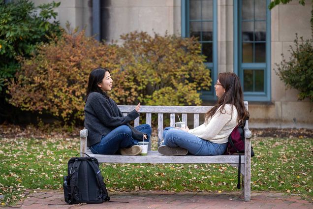 Photo: Two young women sitting cross legged on a park bench, chatting.