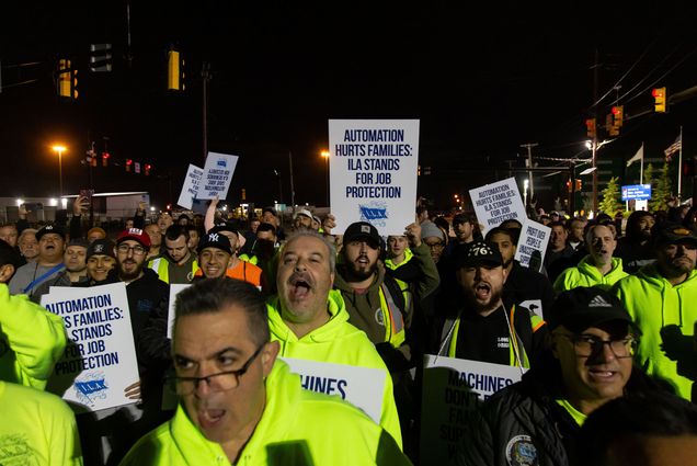 Photo: Dockworkers striking at night. They are wearing safety vests and holding signs that say "Automation Hurts Families: ILA Stands For Job Protection"