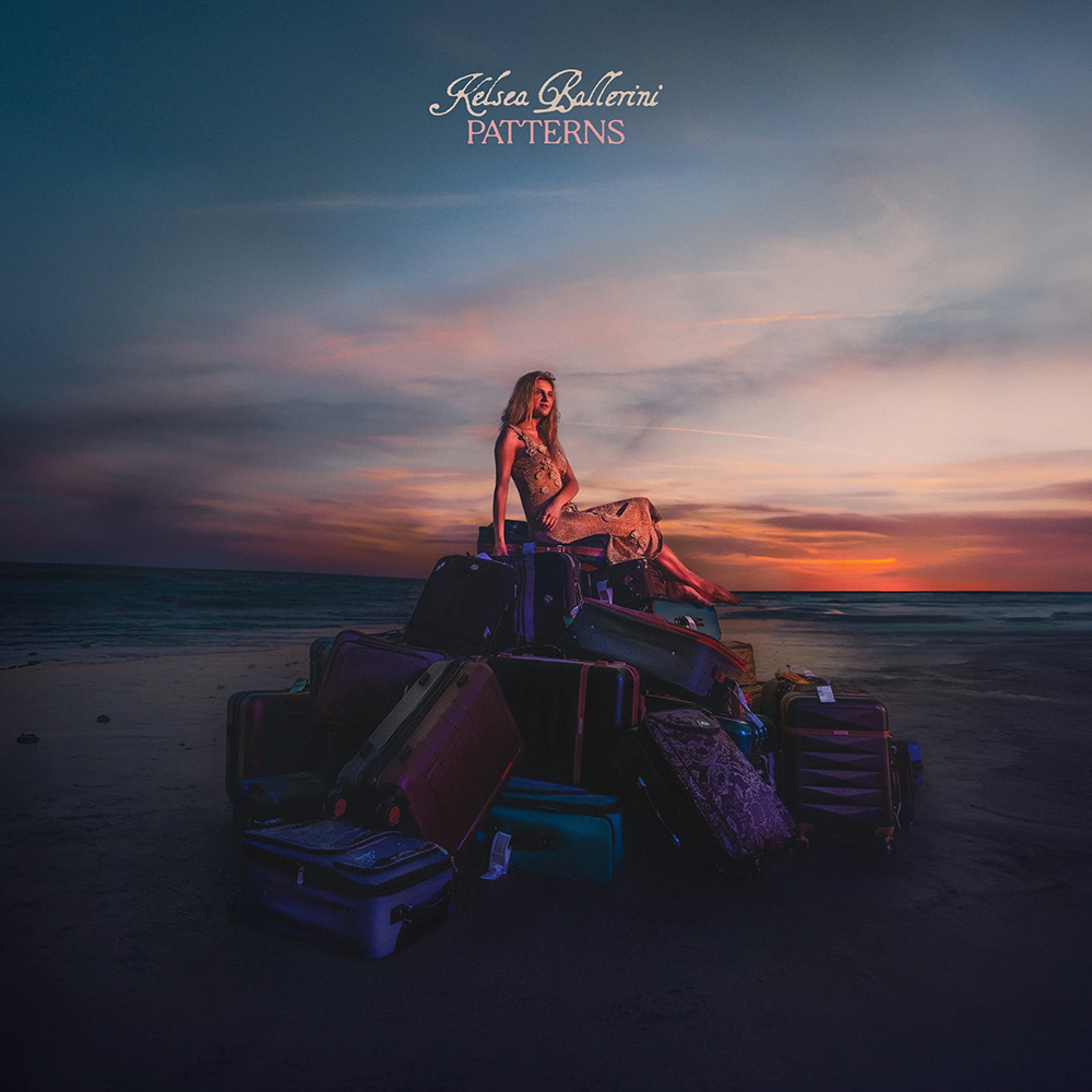 Photo: A women sitting on a huge pile of luggage on a beach. The sun has almost set behind her, casting an orange and blue glow.