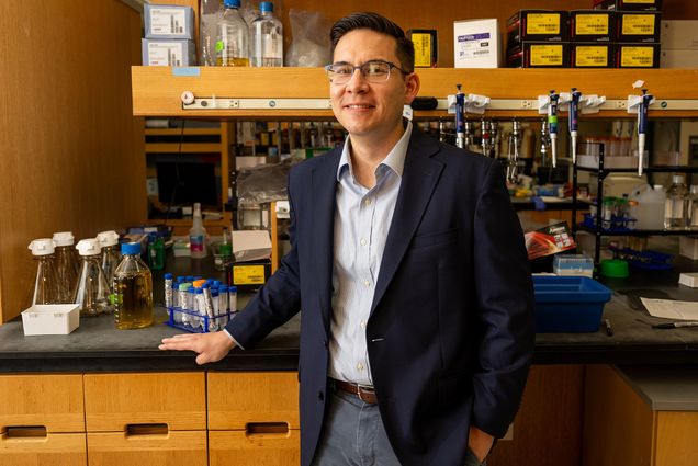 Photo: A picture of a man with short, dark hair and glasses wearing a navy blazer, light blue and white collared button down shirt, a brown belt, and gray pants standing in front of lab equipment.