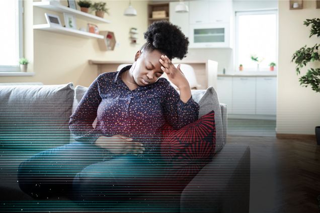 Photo: Young Black woman sitting on a couch with her hands holding her stomach. She looks exasperated. There is a glitch overlay on the image