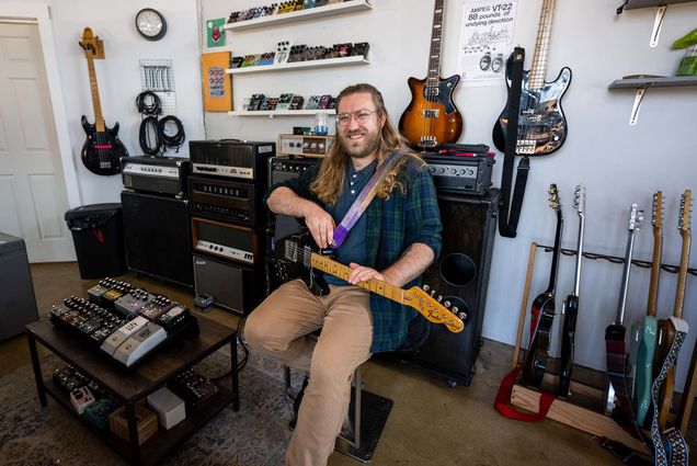 Photo: A picture of a man with long hair wearing a green and blue flannel sittingo n a chair and holding a guitar. He is surrounded by other guitars and guitar pedals