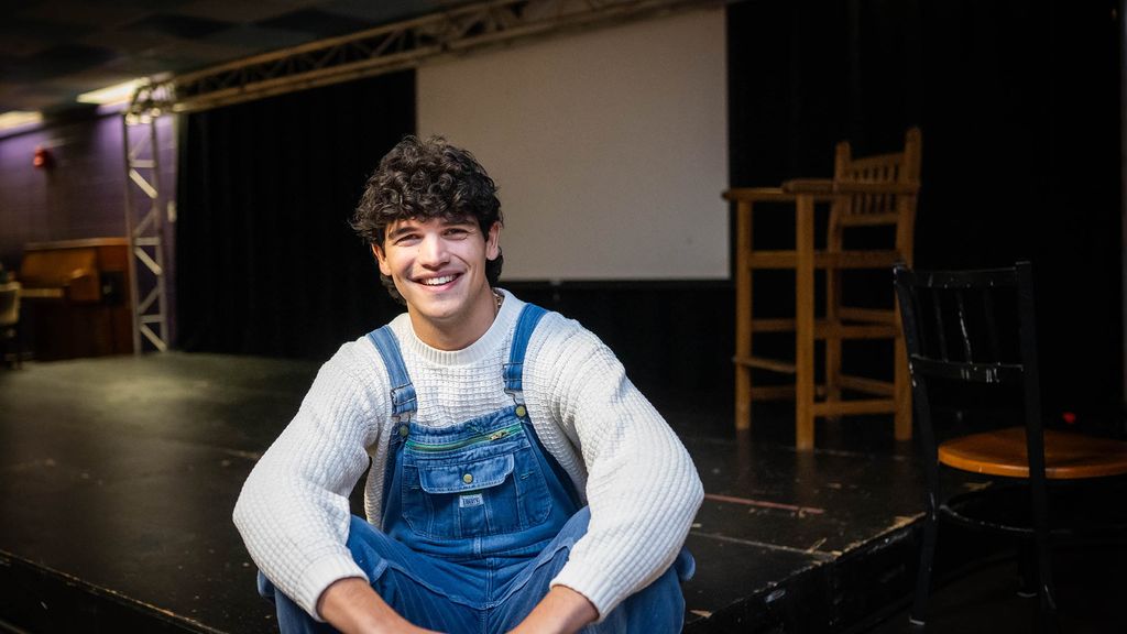 Photo: A young white man with jean overalls and a white shirt smiles while sitting on the ground.