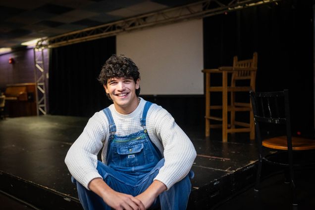 Photo: A young white man with jean overalls and a white shirt smiles while sitting on the ground.