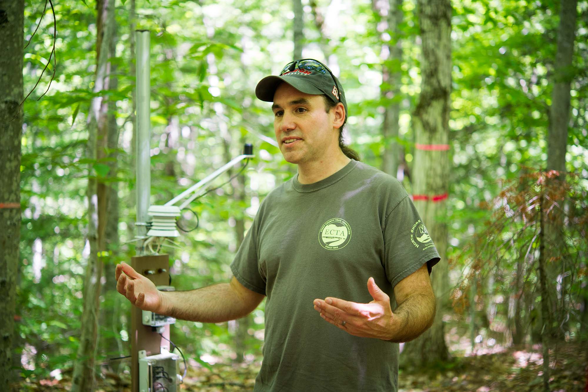 Photo: A man in an olive green baseball cap and t-shirt speaking in the middle of a forest with his arms extended
