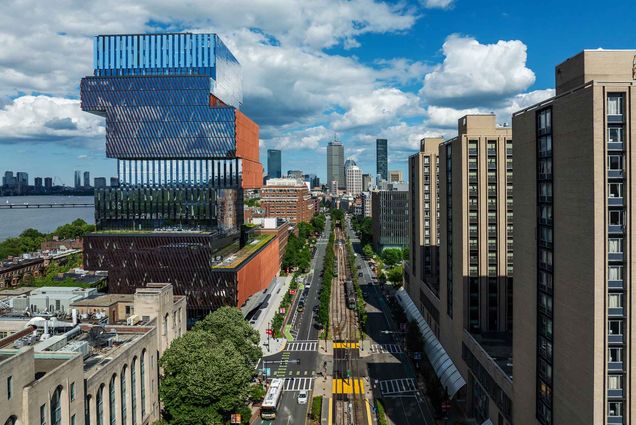 Photo: View of Boston University's urban campus featuring a striking multi-layered glass and metal building, tree-lined streets with tram tracks, and high-rise buildings in the background under a partly cloudy sky.