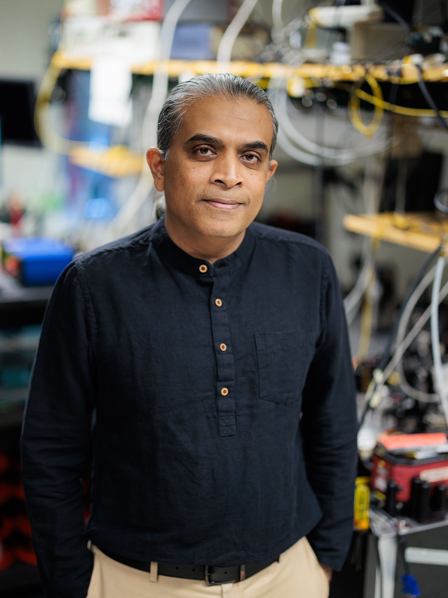 Photo: A man in a dark blue half-button down long-sleeve standing in a lab with his hands in his pockets