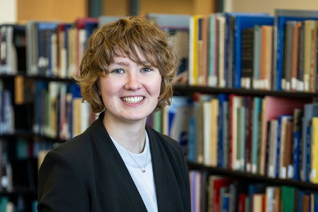 Photo: A white woman with short brown hair poses for a portrait. She sits ontop of a table with a bookcase behind her.
