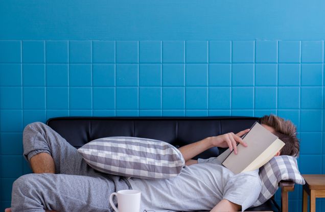 Stock photo of a person with a book lying over their face, while they sleep on a grey couch. There is a pillow laying on top of them, with a mug sitting on the coffee table next to them. Photo credits: Canva.com