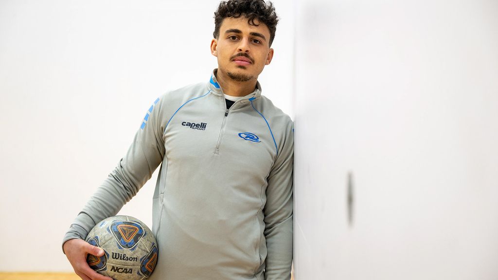 Photo: A young man with short dark hair holding a soccer ball leaning against a wall