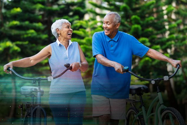 Photo: Two older people with darker complexion walk bikes as they smile and laugh with each other