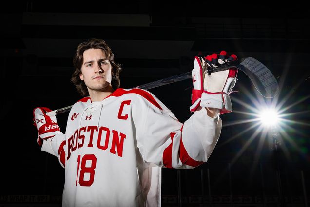 Photo: A hockey player posing in his Boston University uniform with a hockey stick in front of a black background