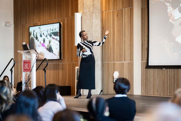 Photo: BU President Melissa Gilliam standing in front of a crowd and speaking. She is wearing a black and white striped shirt, long black skirt, tights, and black pointed-toe heels
