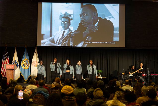 Photo: A group performing in denim jackets on stage in front of a crowd, with a projector screen showing an image of MLK Jr. behind them