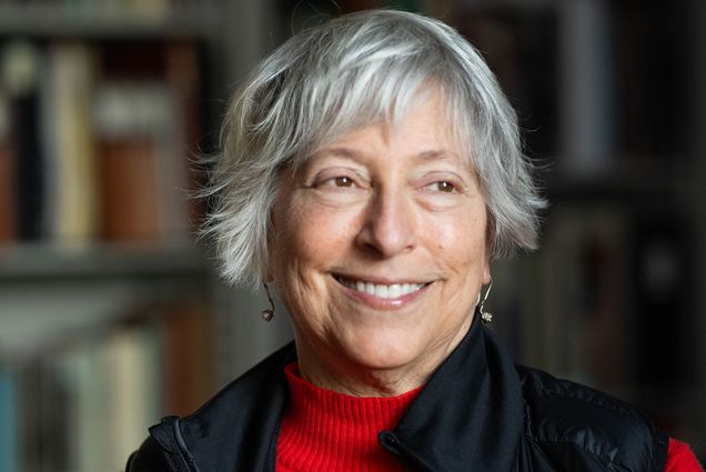 Photo: A woman with gray, short hair smiling and looking off to the side. She is wearing a red turtleneck sweater with a black vest