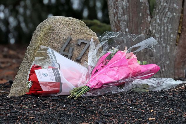 Photo: In the center of the photo, a bouquet of flowers wrapped in plastic and pink paper. They are laying on a dirt ground and are in front of a stone with the number 47 in metal on it. Behind the rock, there is a tree trunk to the right of the photo.