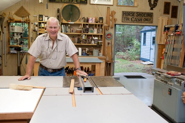 Photo: BU Alum Russell Morash standing behind a table in his home in Lexington, surrounded by tools and other workshop equipment
