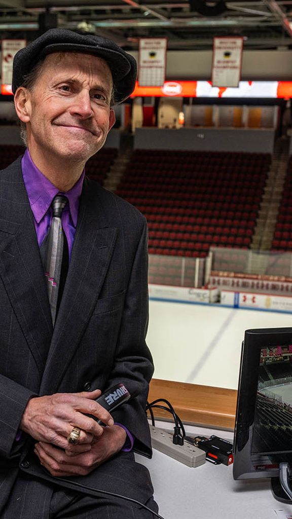 photo: A man wearing a striped black suit, purple shirt, black tie, and black bowler cap while leaning on an announcer's table at the BU hockey arena