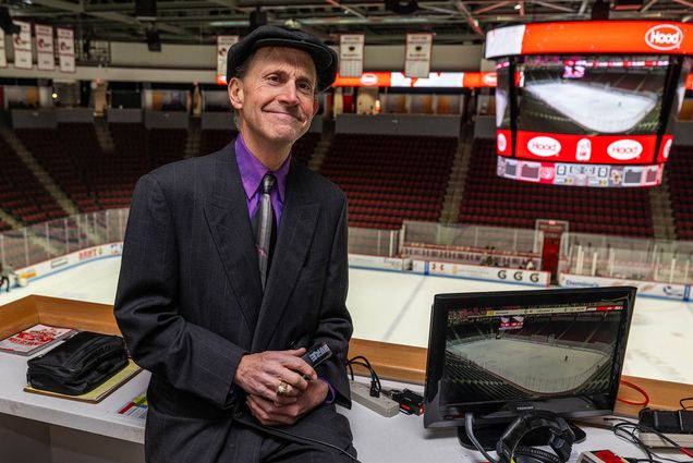 photo: A man wearing a striped black suit, purple shirt, black tie, and black bowler cap while leaning on an announcer's table at the BU hockey arena