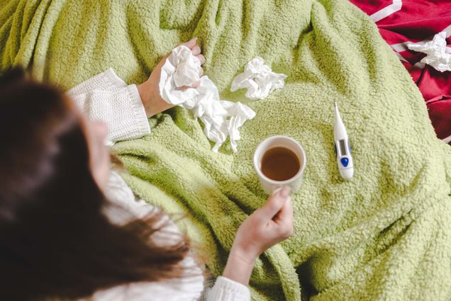 Photo: An above view of a woman sitting with a green blanket wrapped around her, tea in her hand, and crumpled up tissues strewn about the blanket