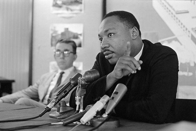 Photo: A black-and-white picture of Martin Luther King Jr. sitting down and speaking into a microphone with a man sitting to his left in the background