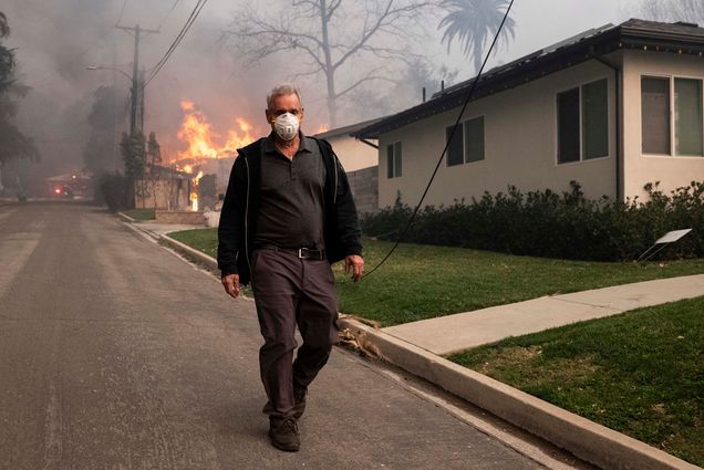 Photo: A man with a mask walks on the road, flames and smoke blazing behind him.