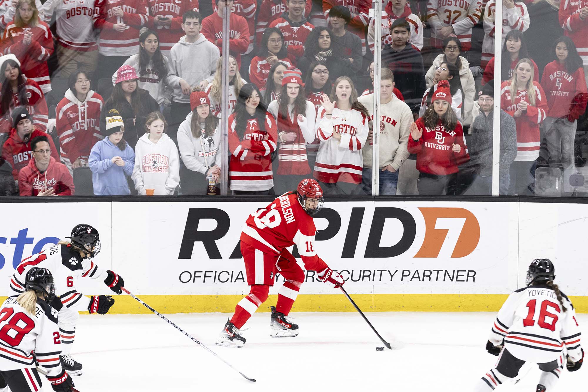 Photo: A BU women's hockey player shuffling the puck as a crowd of BU supports watch from the stands.