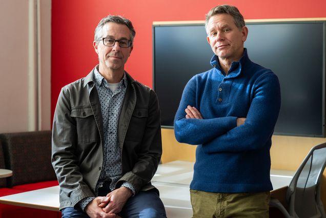 Photo: Two men posing next to each other. The one on the left has gray hair, glasses, a dark jacket, and a patterned shirt. The one on the right has gray hair, a blue sweater with buttons at the top, and khakis