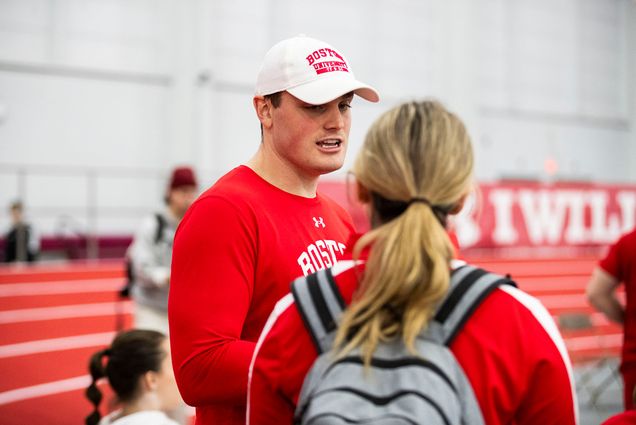 Photo: A man wearing a white Boston University ballcap speaks with another individual at an indoor track