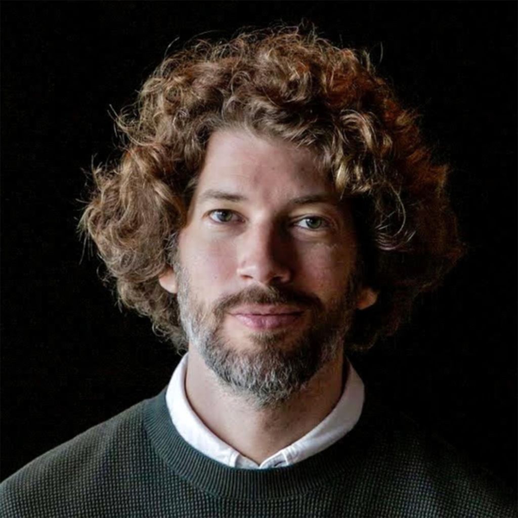 Photo: A headshot of a man with curly brown hair and a beard wearing a sweater with a white collared shirt under it