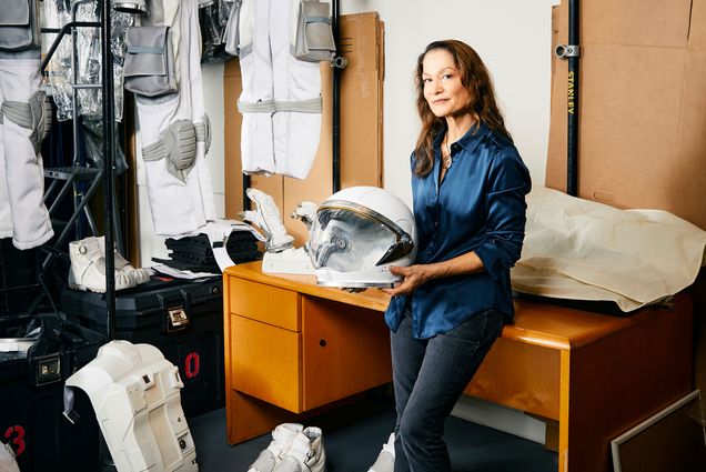 Photo: A woman leaning against a desk as she holds an astronaut helmet. The wall behind her has astronaut suits across it and a poster with an astronaut that says "Apply Now"