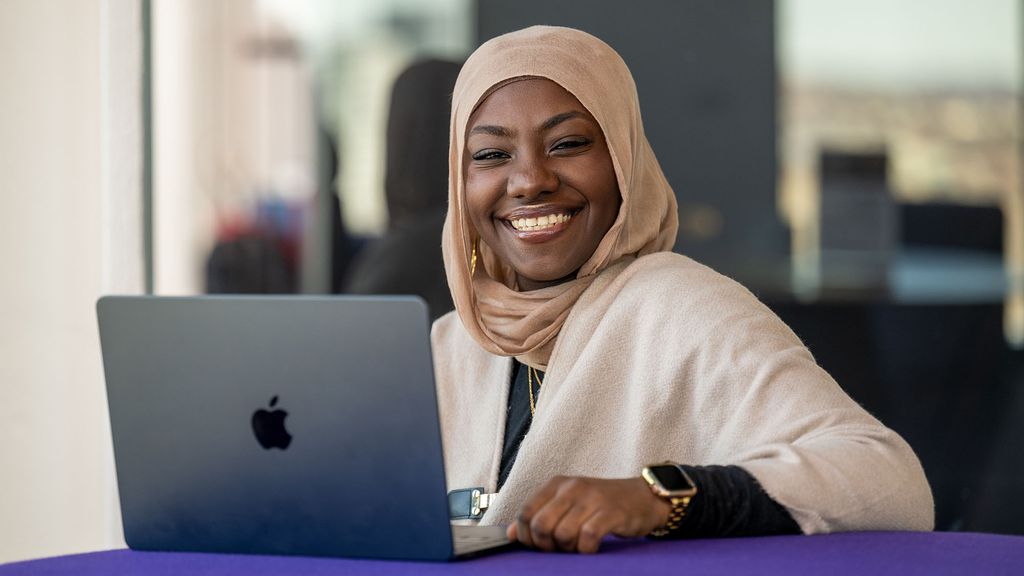Photo: A Black woman wearing a hijab smiling and sitting behind an open laptop.