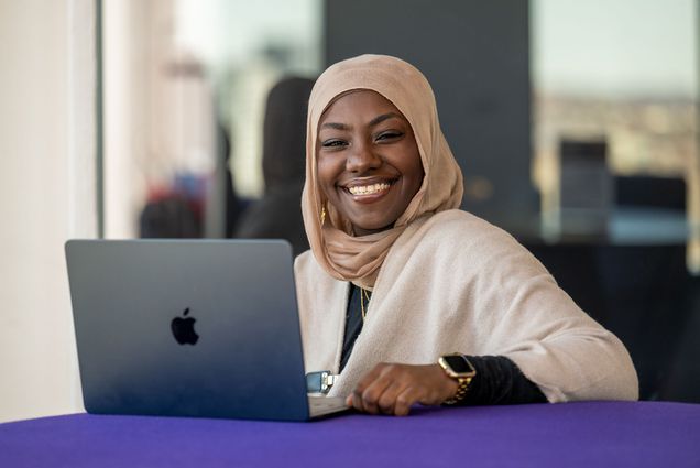 Photo: A Black woman wearing a hijab smiling and sitting behind an open laptop.