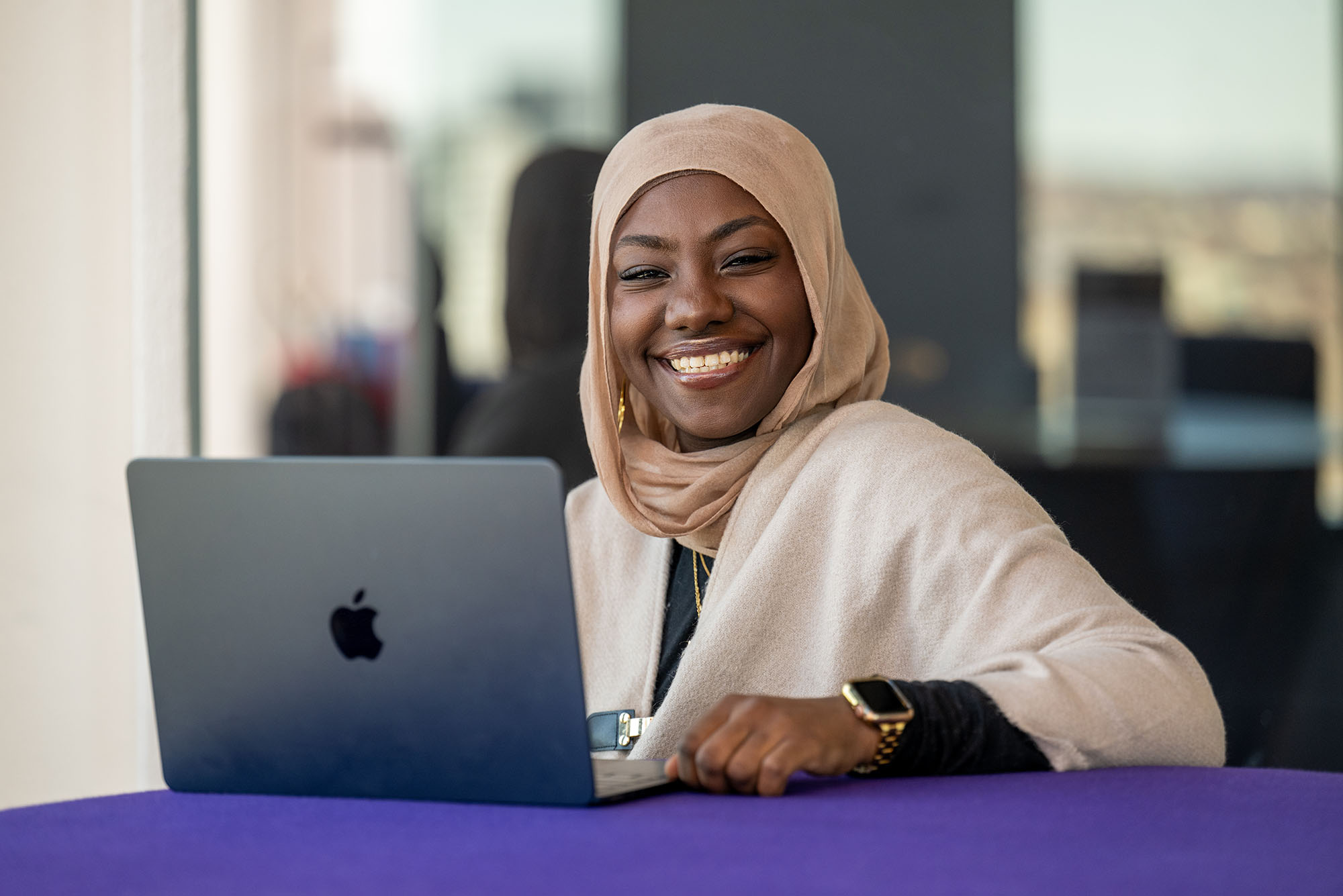 Photo: A Black woman wearing a hijab smiling and sitting behind an open laptop.