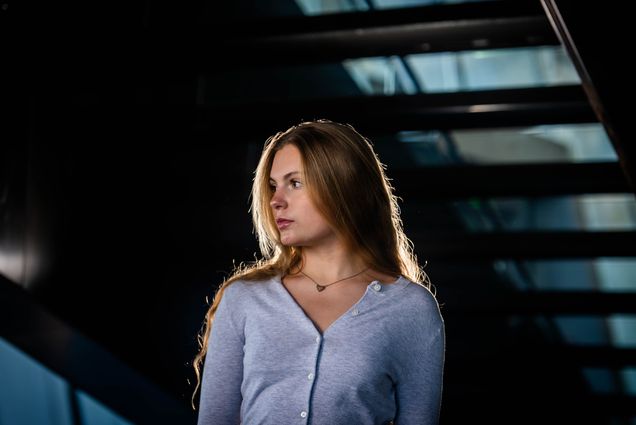 Photo: A blonde female student wearing a gray cardigan and looking off to the side as she stands under a staircase