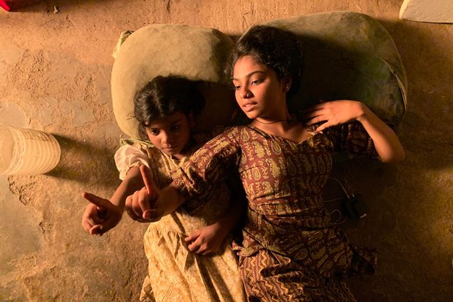Photo: A still from the short film, Anuja. Two young women sit on a cot and point to the left.