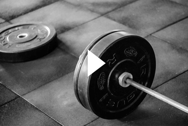 Photo: Stock image of a barbell on a gym mat. A play button overlay rest on top the image.