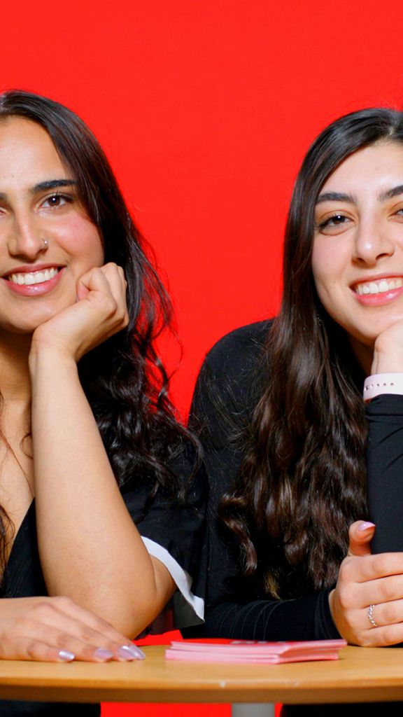 Photo: Two girls smiling and posing with their head resting against their hand as they sit behind a table. There is a red background behind them