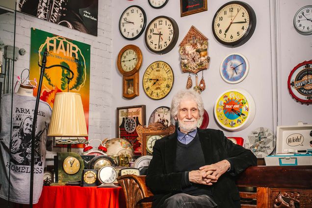 Photo: A distinguished man with white hair sits with hands folded in front of a wall of ornate clocks and other items