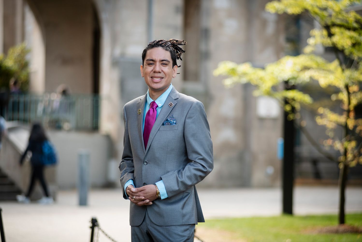 Photo: An individual with long dark hair smiling in a grey suit outdoors on a sunny day