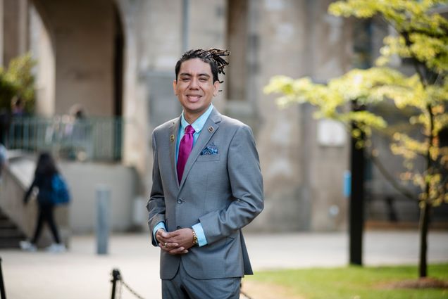 Photo: An individual with long dark hair smiling in a grey suit outdoors on a sunny day