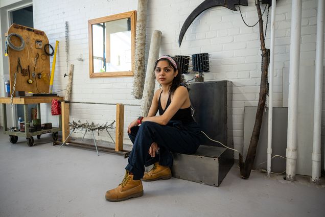 Photo: A woman with long dark hair wearing a bandana sits on a piece of metal inside of an art studio