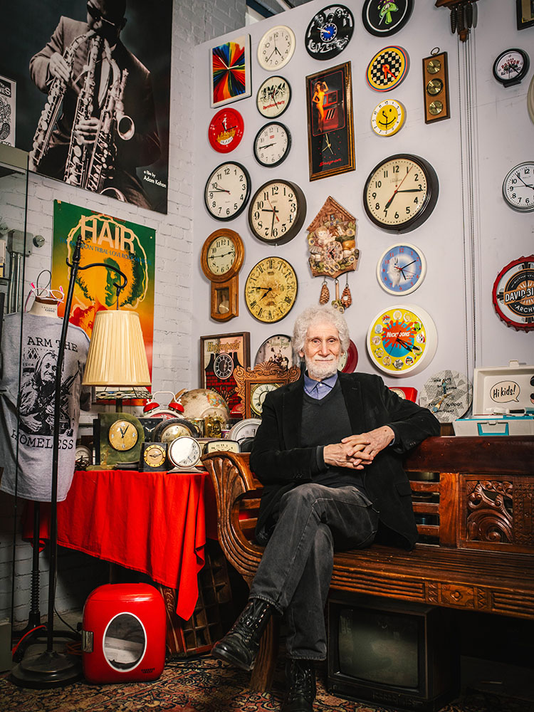 Photo: A distinguished man with white hair sits with hands folded in front of a wall of ornate clocks and other items