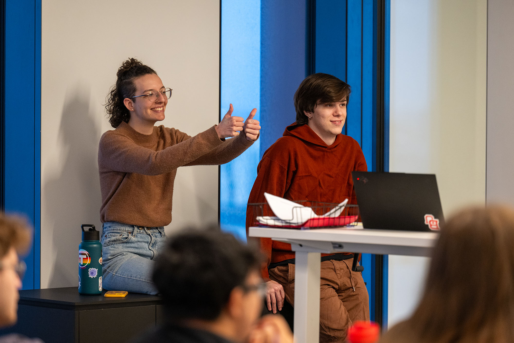 Photo: Two people sitting in front of a larger group, one on the left is giving a thumbs up and the other sits in front of a computer