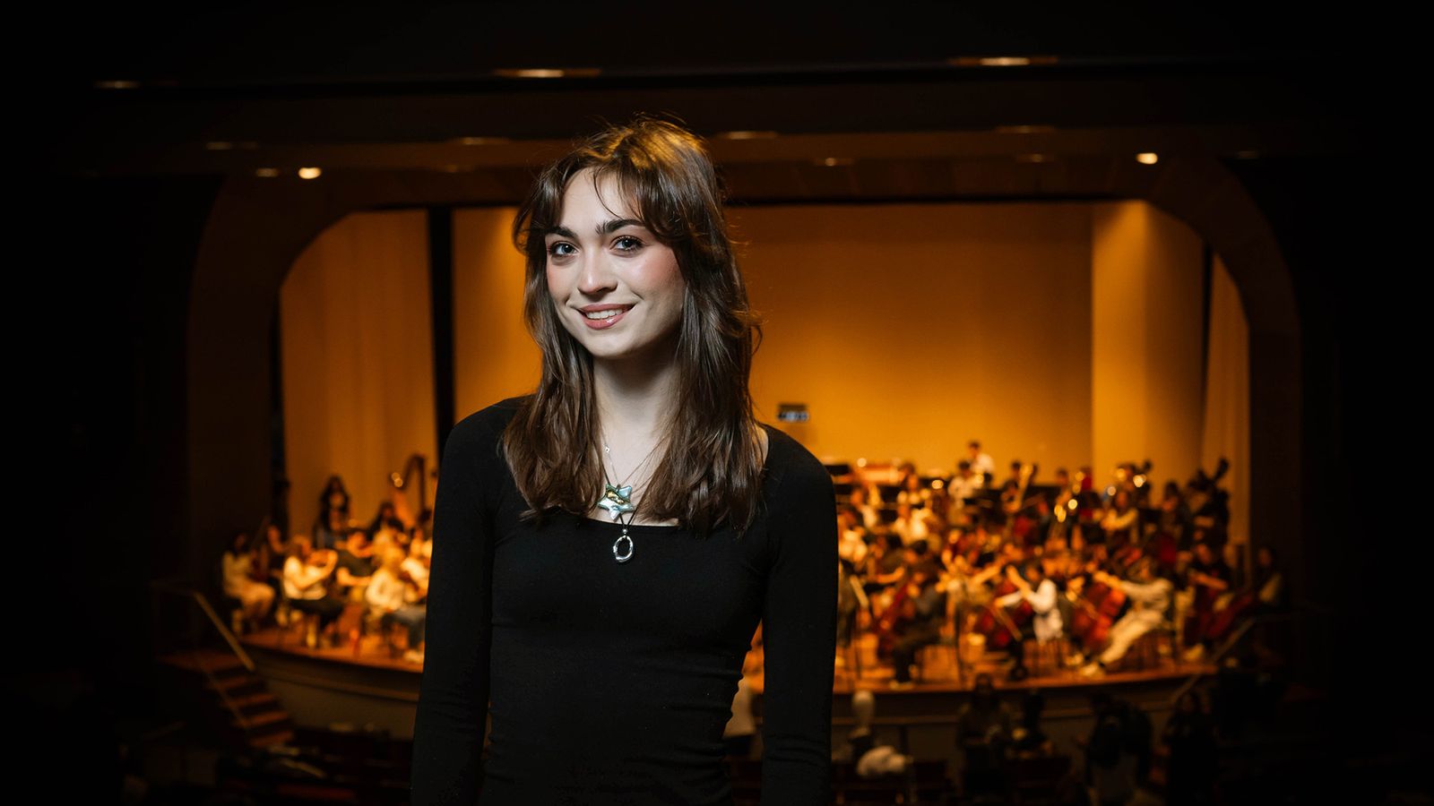 Photo: A girl with brown hair and bangs wearing a long sleeve black shirt and smiling in front of a stage
