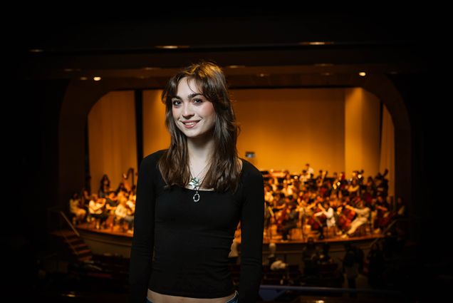 Photo: A girl with brown hair and bangs wearing a long sleeve black shirt and smiling in front of a stage